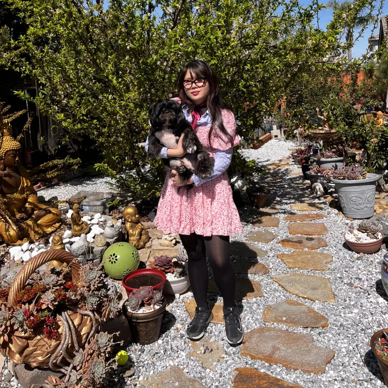 Woman in neckerchief with blue polka dot dress shirt underneath pink floral dress with black and white shih tzu