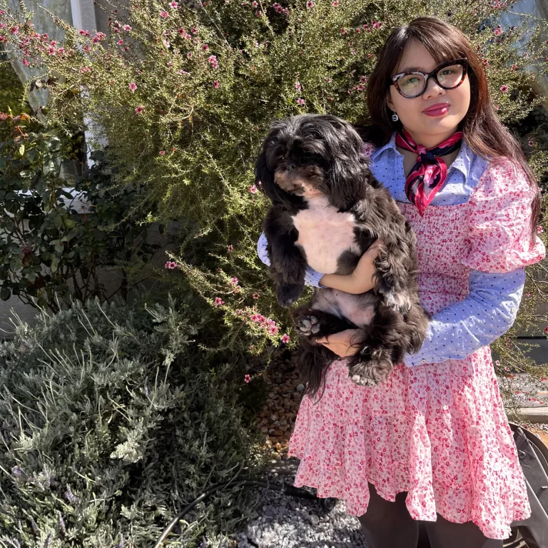 Woman in neckerchief with blue polka dot dress shirt underneath pink floral dress with black and white shih tzu